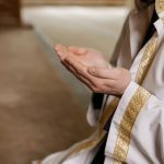 A man in traditional Middle Eastern attire prays indoors, hands clasped together.